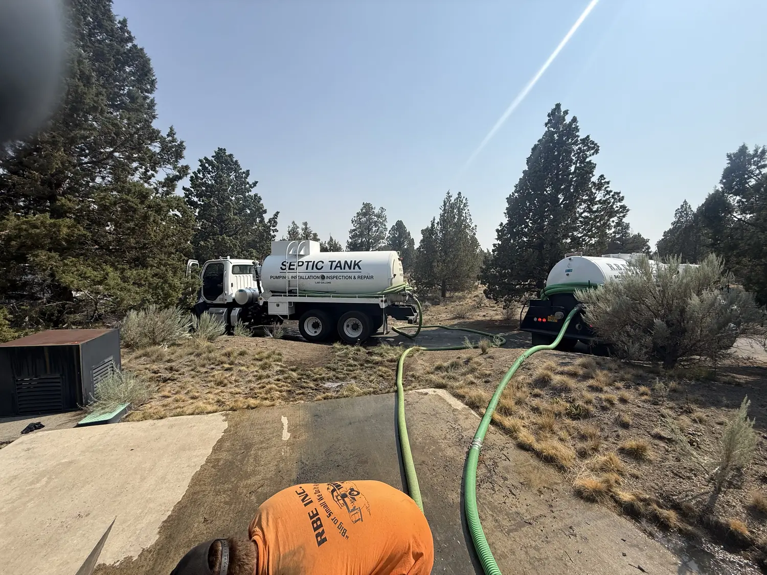 Two RBE Inc. septic pump trucks parked among junipers with hoses run to a tank during a pump-out near Bend, Oregon